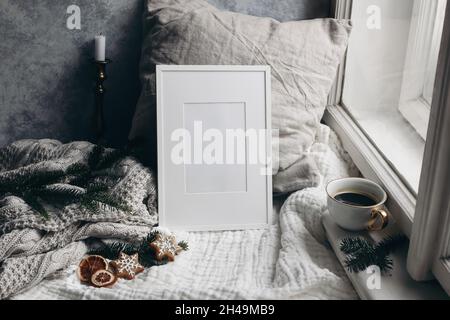Christmas still life. Candle in golden candleholder. White picture frame mockup. linen pillow near window. Moody composition. Fir tree branches, cup Stock Photo