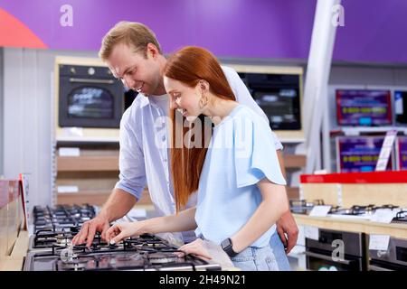 Nice Couple Buying Electrical Oven With Consumer Loan Credit, In Store, Examining Oven Stove, Discussing. Married Couple Buying Household Appliances I Stock Photo