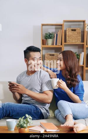 Positive young Asian couple looking at each other and enjoying time at home: man using smartphone, his girlfriend drinking coffee Stock Photo