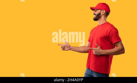 Male Delivery Worker In Red Uniform Pointing Finger Aside Showing Empty Space For Advertisement Text, Advertising Courier Service Posing Over Yellow S Stock Photo