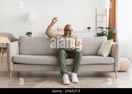 Mature African American Man Using Laptop Pointing Remote Controller Up Turning On Air Conditioner Sitting On Couch In Living Room At Home. Domestic Co Stock Photo