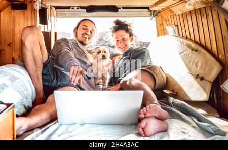 Independent couple with cute dog using laptop on retro mini van transport - Travel life inspiration concept with indie man and woman on minivan Stock Photo
