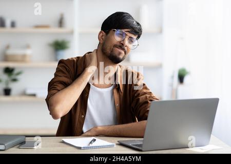 Irritated young arabic man in glasses suffering from office syndrome, sitting at worktable in front of modern laptop, touching his neck, having pain, Stock Photo
