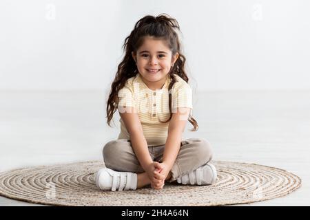 Portrait Of Cute Smiling Little Girl Sitting With Legs Crossed At Wicker Carpet At Home, Beautiful Cheerful Adorable Female Child Relaxing On Floor, L Stock Photo