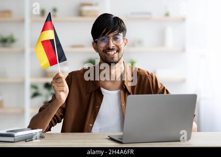 Smiling indian guy student sitting at table at home, showing flag of Germany, using laptop, copy space. Positive handsome young man studying German on Stock Photo
