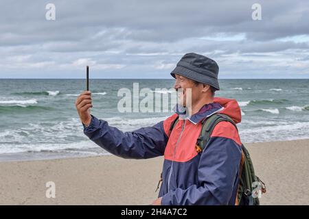 Senior asian man with backpack on seashore talking via video communication. Stock Photo