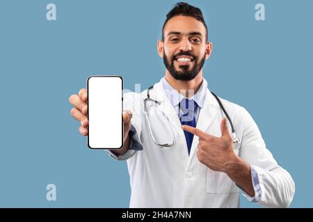 Middle Eastern Male Doctor Showing Empty Cellphone Screen Advertising Clinic Website Or Medical App Standing Over Blue Studio Background, Smiling To C Stock Photo