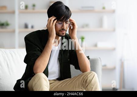 Cheerless indian guy in casual with closed eyes wearing glasses touching his head, having migraine, suffering from headache or experiencing difficulti Stock Photo