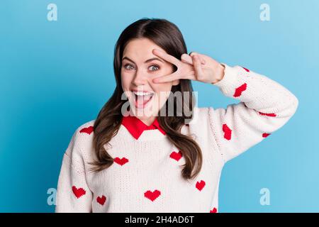Photo of tricky lovely young lady wear hearts print sweater open mouth showing v-sign eye isolated blue color background Stock Photo