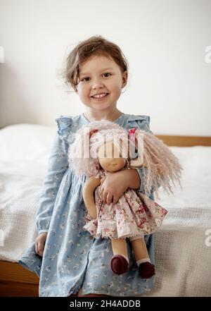 Cute little girl with blond hair holding her doll and smiling Stock Photo