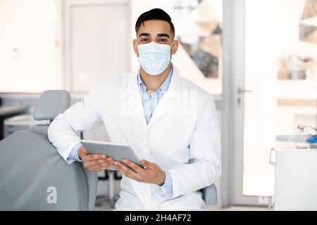 Young Arab Stomatologist Doctor Using Digital Tablet At Workplace In Clinic, Middle Eastern Dentist Wearing protective Medical Mask And Uniform Posing Stock Photo