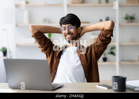 Wealthy indian guy project manager sitting at worktable in front of modern laptop, leaning back and smiling, feeling good about his job, office interi Stock Photo