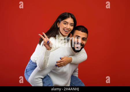 Joyful arab couple having fun and showing peace sign, happy guy piggybacking his wife and smiling at camera while posing together over red background, Stock Photo