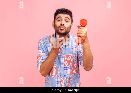 Young adult romantic man call operator wearing blue casual style shirt holding red landline phone and sending air kissing to camera. Indoor studio shot isolated on pink background. Stock Photo