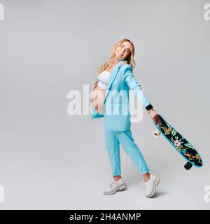 a pregnant girl in turquoise clothes with a skateboard in her hands on a gray background. Stock Photo