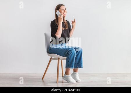 Smiling young woman having conversation on cellphone, sitting on cozy chair against white studio wall, full length. Beautiful Caucasian lady talking w Stock Photo