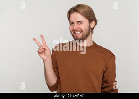 Portrait of man with beard wearing sweatshirt, doing victory gesture and smiling to camera, showing peace, v sign with double fingers. Indoor studio shot isolated on gray background. Stock Photo