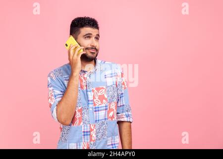 Young adult man with beard in blue casual style shirt talking phone, calling to discuss plans, looking away with dreamy facial expression. Indoor studio shot isolated on pink background. Stock Photo
