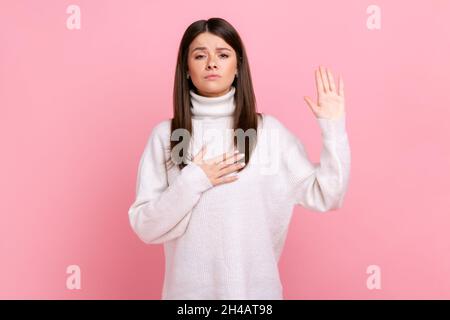 Portrait of honest young woman standing with hand on chest and fingers up, making loyalty promise, wearing white casual style sweater. Indoor studio shot isolated on pink background. Stock Photo