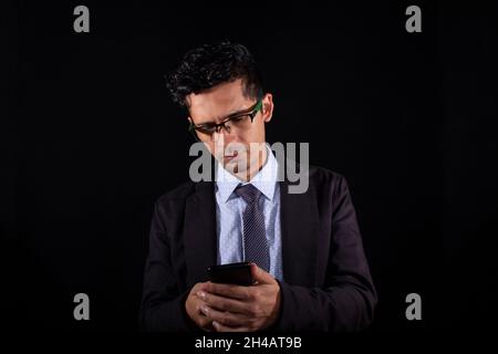 Business man looking at his smart phone, isolated on black background. Young adult business man wearing glasses for looking at his smart phone. Latin Stock Photo