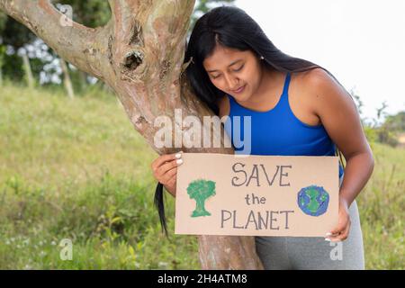 A brunette woman holds a sign calling to save the planet as she reads it. Latina girl protects nature and trees. Stock Photo