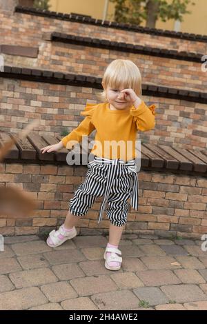 A beautiful cheerful little girl in a stylish striped autumn suit is playing near a bench in a city park. Lifestyle Stock Photo