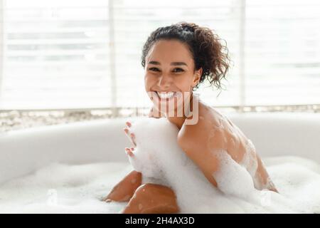 Portrait of cheerful young woman taking bath with foam, smiling at camera, relaxing in hot bathtub in morning at home. Lovely millennial female enjoyi Stock Photo