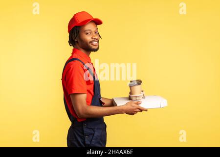 Side view portrait of optimistic professional courier standing with pizza box and coffee in disposable cup, fast food delivery service. Indoor studio shot isolated on yellow background. Stock Photo