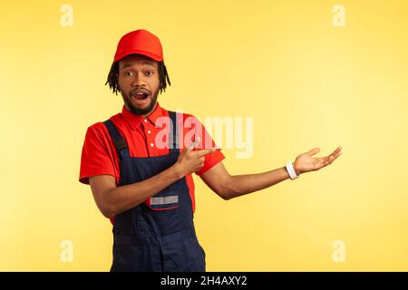 Astonished builder or courier wearing overalls, presenting copy space for advertisement, pointing finger aside, ad of delivery service. Indoor studio shot isolated on yellow background. Stock Photo