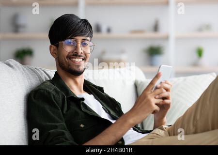Positive arabic guy playing mobile games on cellphone while reclining on sofa at home, looking at camera and smiling, enjoying newest mobile applicati Stock Photo