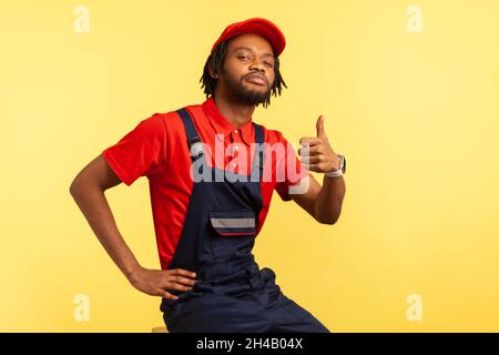 Portrait of handsome confident handyman wearing blue uniform, red visor cap and T-shirt posing and showing thumbs up, looking at camera. Indoor studio shot isolated on yellow background. Stock Photo