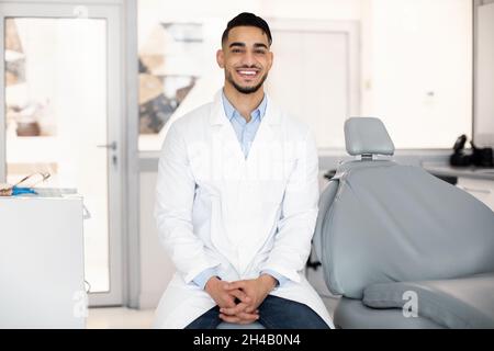 Stomatological Services. Handsome Arab Male Dentist Posing At Workplace In Clinic Interior, Middle Eastern Stomatologist Doctor In Uniform Looking And Stock Photo