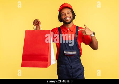 Cheerful positive delivery man in uniform pointing finger at shopping bags in his hand, fast delivery from store, service industry. Indoor studio shot isolated on yellow background. Stock Photo