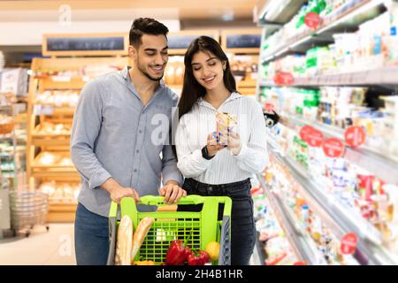 Middle Eastern Spouses Holding Dairy Products Doing Grocery Shopping Together In Modern Supermarket. Arab Family Buying Food In Store On Weekend. Cons Stock Photo