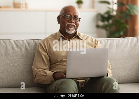 Happy Mature Black Man Using Laptop Browsing Internet Smiling To Camera Working Online Sitting On Sofa At Home, Wearing Glasses. Freelance In Older Ag Stock Photo