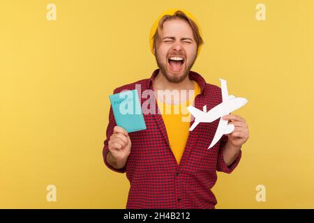 Portrait of crazy excited hipster bearded guy in beanie hat and checkered shirt holding passport document and airplane mockup, rejoicing travel tour. Indoor studio shot isolated on yellow background. Stock Photo