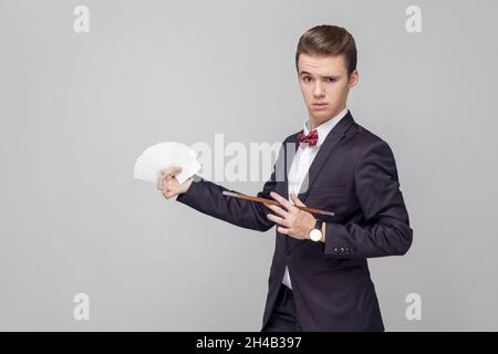 Portrait of elegant magician with stylish hairdo in black tuxedo holding magic wand and pointing at playing cards in his hands, showing trick, illusionist performance. studio shot, gray background Stock Photo
