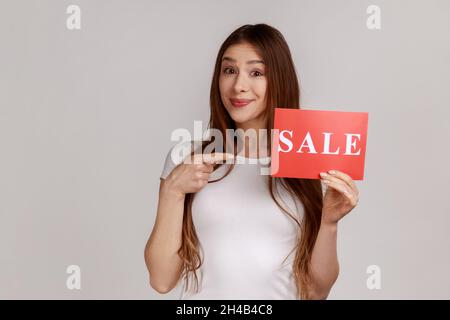 Portrait of beautiful dark haired woman holding pointing at sale word, looking smiling at camera, presenting discounts, wearing white T-shirt. Indoor studio shot isolated on gray background. Stock Photo