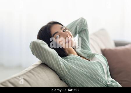 Lazy weekend at home. Charming Indian woman resting with hands behind head on cozy couch at living room. Millennial Eastern lady relaxing on comfortab Stock Photo