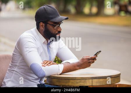 The young man is sitting at a table in a street cafe and watching in the smartphone. He is wearing a fashion white shirt and cap Stock Photo