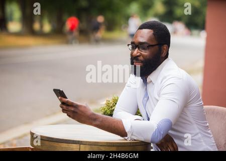 The stylish young man is sitting in a street cafe and uses a smartphone. He straightens his hair while have a video call Stock Photo