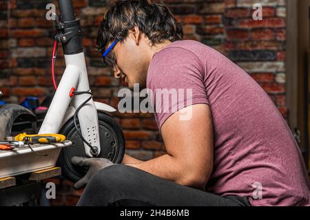 A seated latin mechanic puts a wheel of an electric scooter back in place in his workshop. Stock Photo
