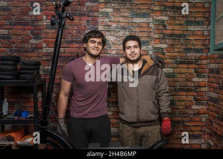 Front view of two mechanics colleagues hugging and looking at the camera standing against a brick wall in the workshop. Stock Photo