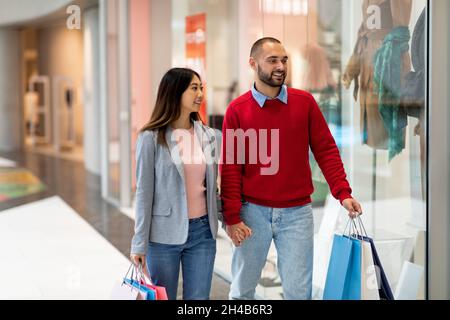 Loving interracial couple walking along store windows, holding hands, shopping for clothes at mall, copy space. Millennial diverse spouses with paper Stock Photo
