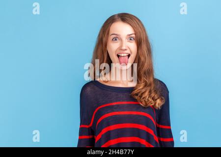 Portrait of funny disobedient young woman wearing striped casual style sweater demonstrating tongue, behaving naughty unruly, childish mood. Indoor studio shot isolated on blue background. Stock Photo