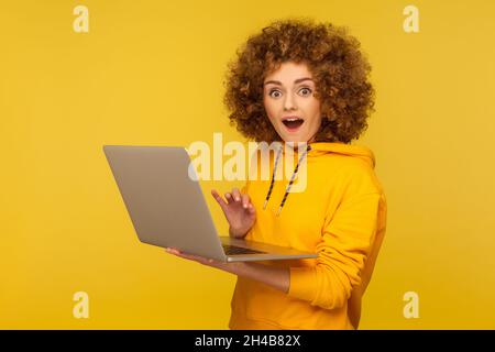 Amazed, surprised woman with Afro hairstyle holding laptop and looking with shocked expression, working on computer, wearing casual style hoodie. Indoor studio shot isolated on yellow background. Stock Photo