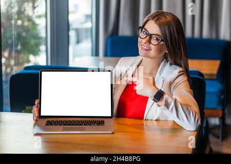 Smiling happy young woman employee in glasses pointing empty laptop screen, mock up display for advertisement, online service, website promotion. Indoor shot, cafe or office background. Stock Photo