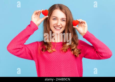 Portrait of optimistic young adult woman wearing pink pullover, having fun with red toy hearts, showing his fondness and devotion, romance. Indoor studio shot isolated on blue background. Stock Photo