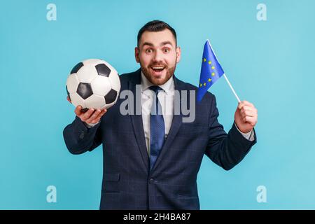 Excited man wearing official style suit, holding black and white soccer ball and flag of EU, watching football and support favourite team. Indoor studio shot isolated on blue background. Stock Photo