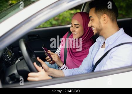 Young Muslim Spouses Riding Car And Using Smartphone Together, Happy Middle Eastern Couple Browsing Online Maps Or Gps Navigation On Mobile Phone, Pla Stock Photo